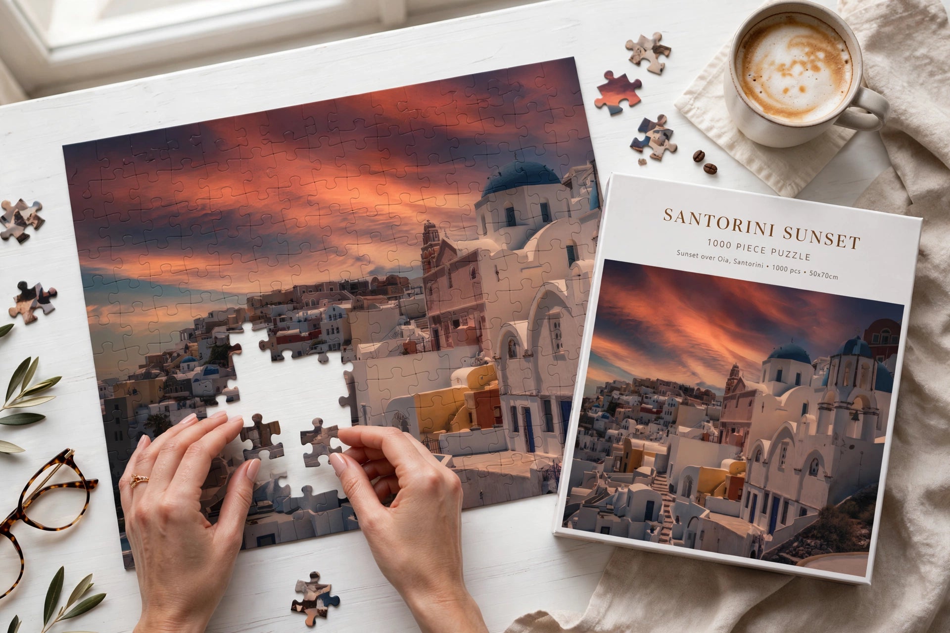 A person's hands are assembling a large jigsaw puzzle of the Santorini Sunset, with the completed puzzle and its box displayed on a white tablecloth alongside a coffee cup, glasses, and olive branches.