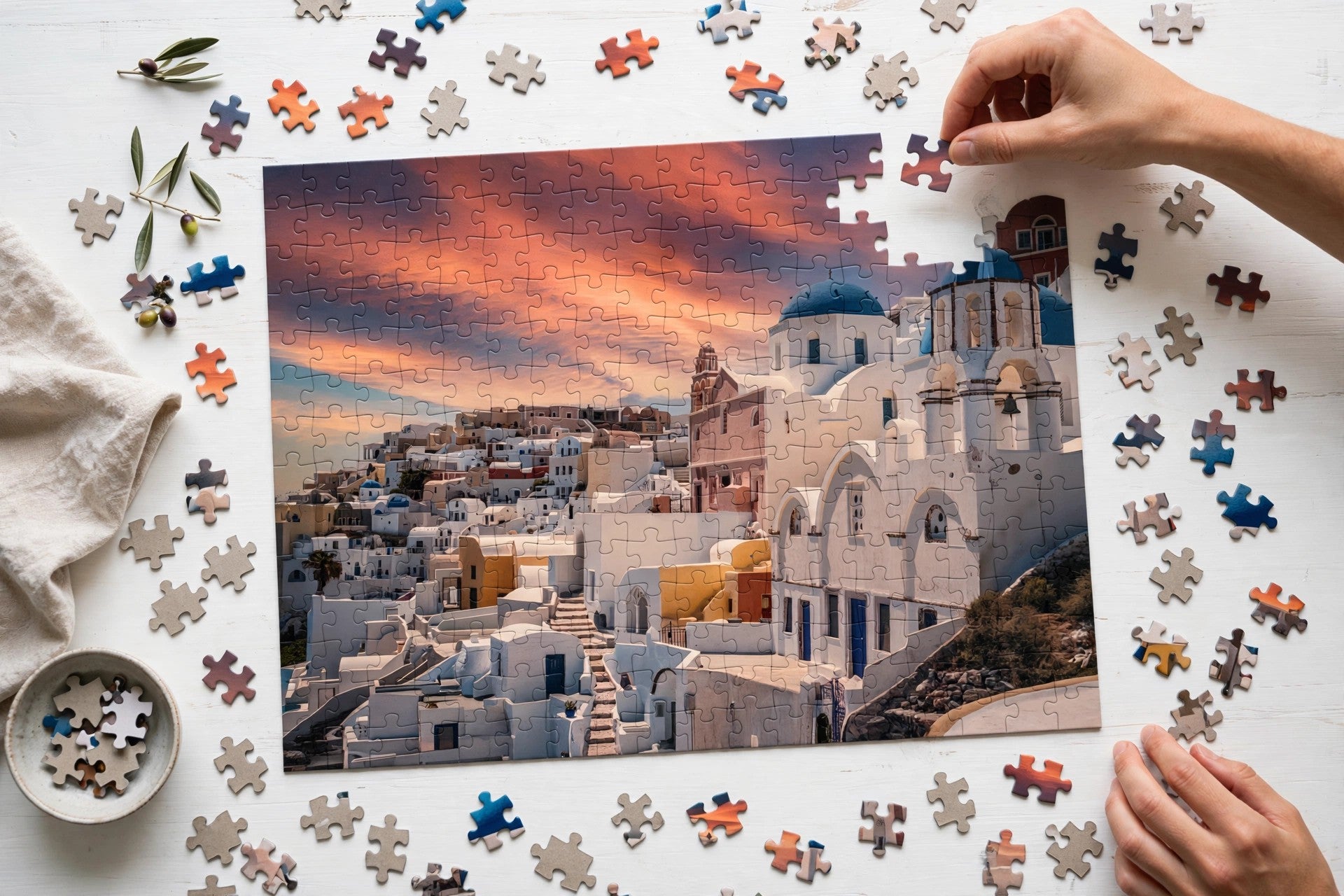 A person's hands are assembling a jigsaw puzzle of a Greek village at sunset, with loose puzzle pieces scattered on a white wooden table.